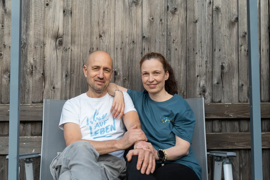 Mentaltrainer Marcus und Verena Bernhardt Österreich sitzen entspannt vor einer Holzwand für Outdoor Coaching in der Nat